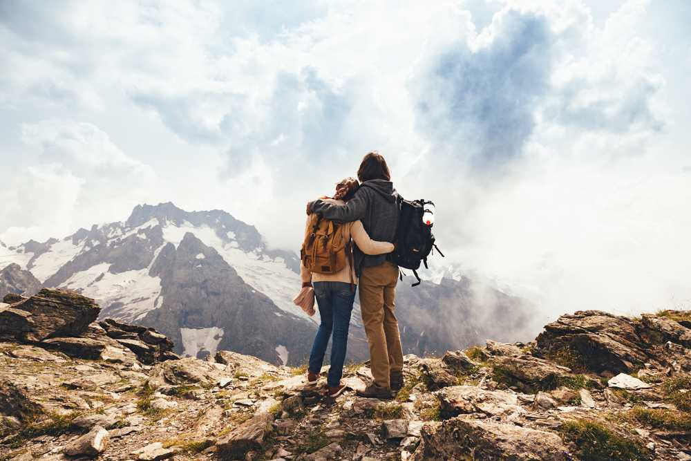 Man and woman with hiking backpacks standing on a rocky summit overlooking snow-capped mountains